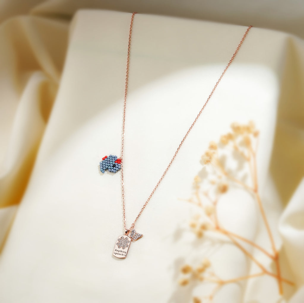 Blue beaded elephant charm and crystal tag pendant on a rose-gold chain over cream fabric