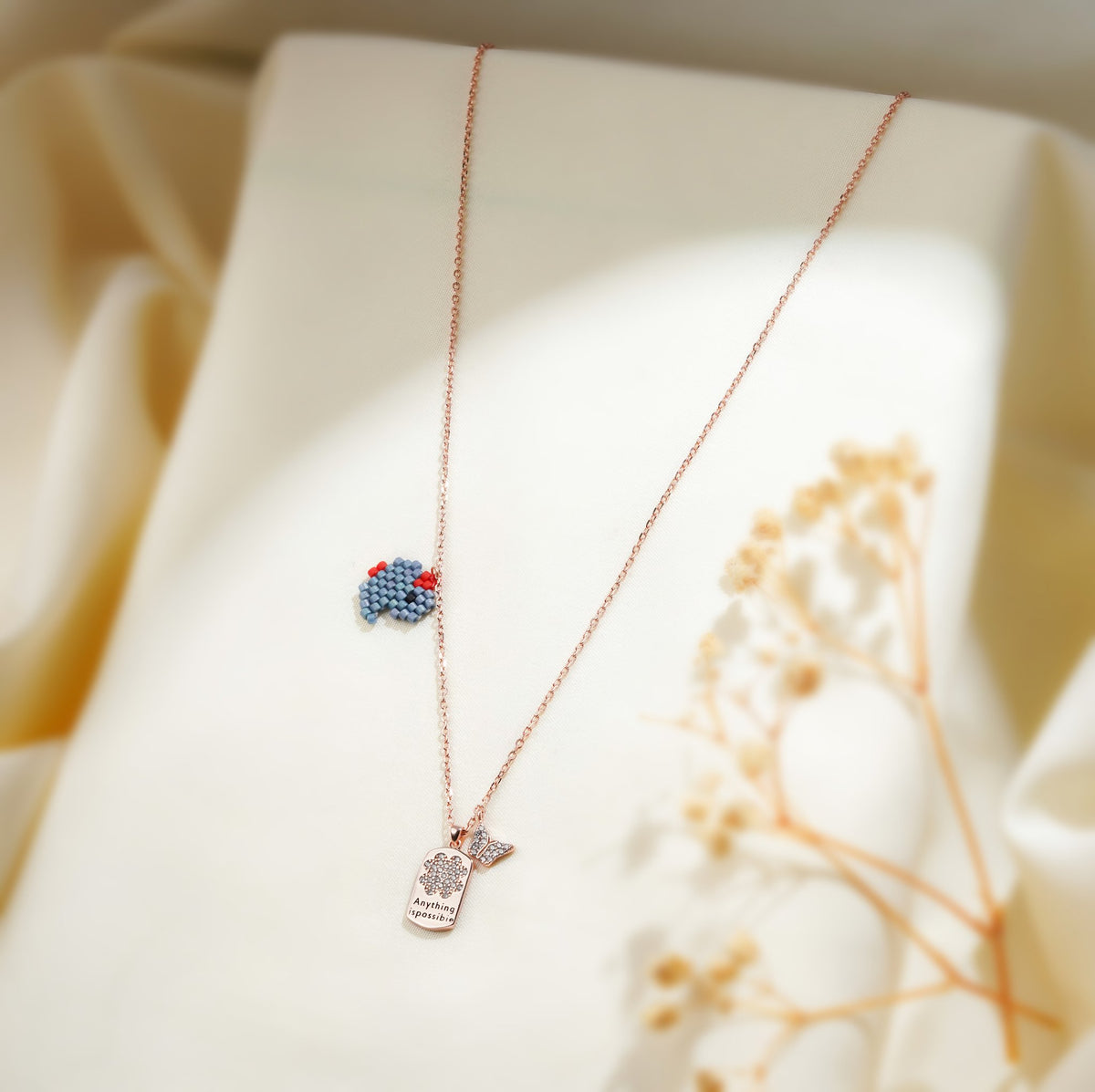 Blue beaded elephant charm and crystal tag pendant on a rose-gold chain over cream fabric
