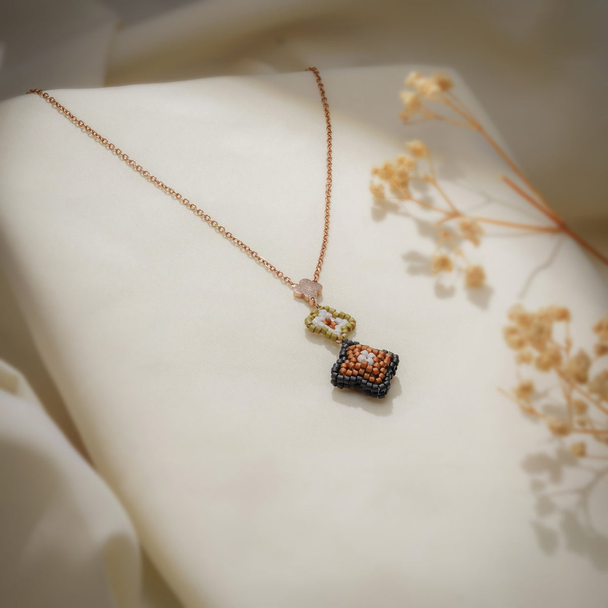 Bronze beaded clover pendant on a rose-gold chain draped over cream fabric