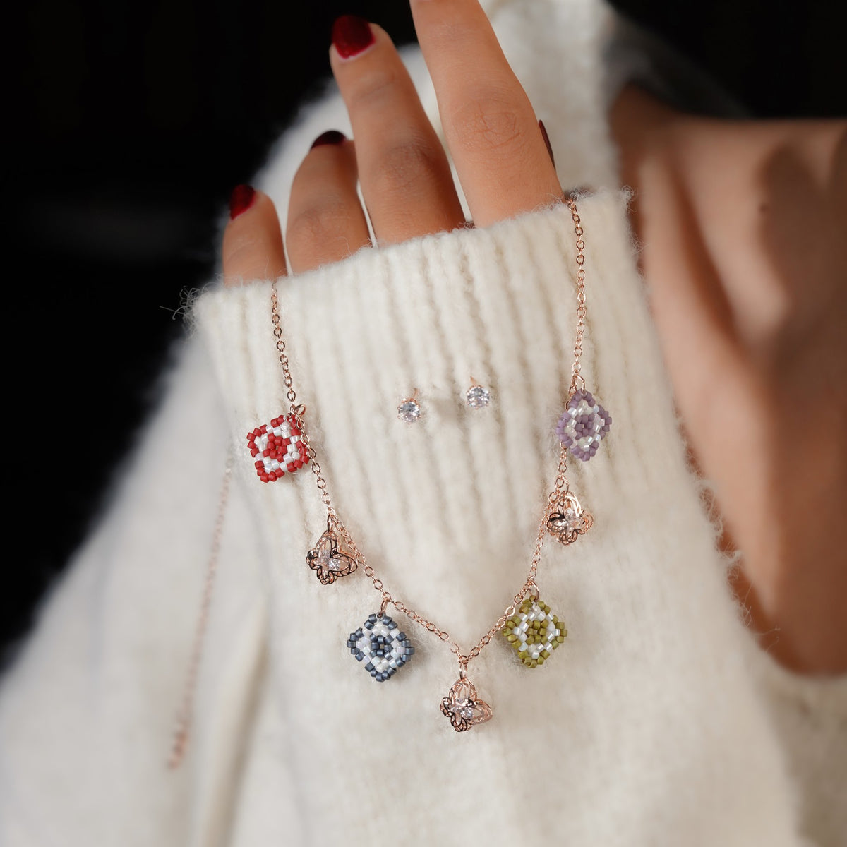 Hand holding the colourful beaded square charm necklace beside a pair of crystal stud earrings