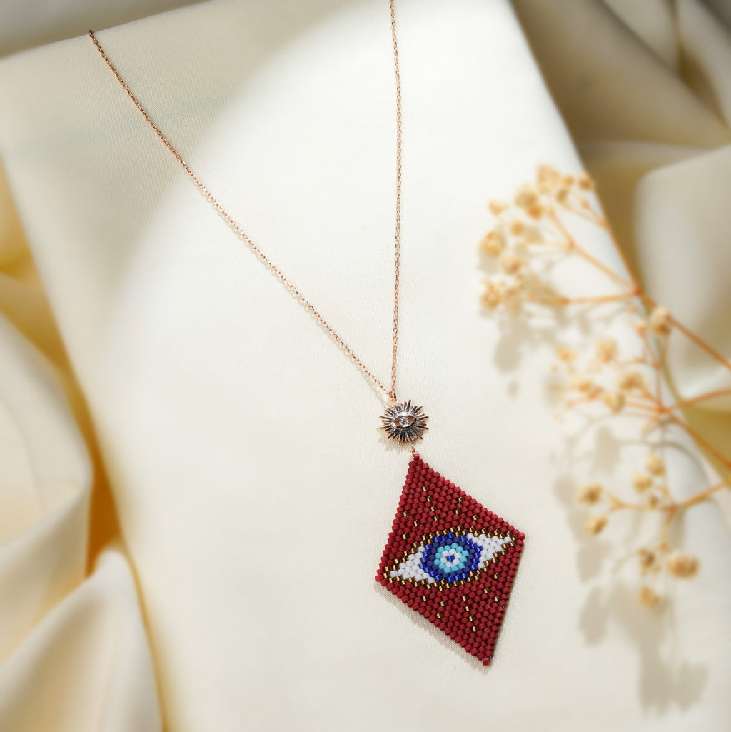 Crimson beaded evil-eye pendant draped on cream fabric beside dried gypsophila