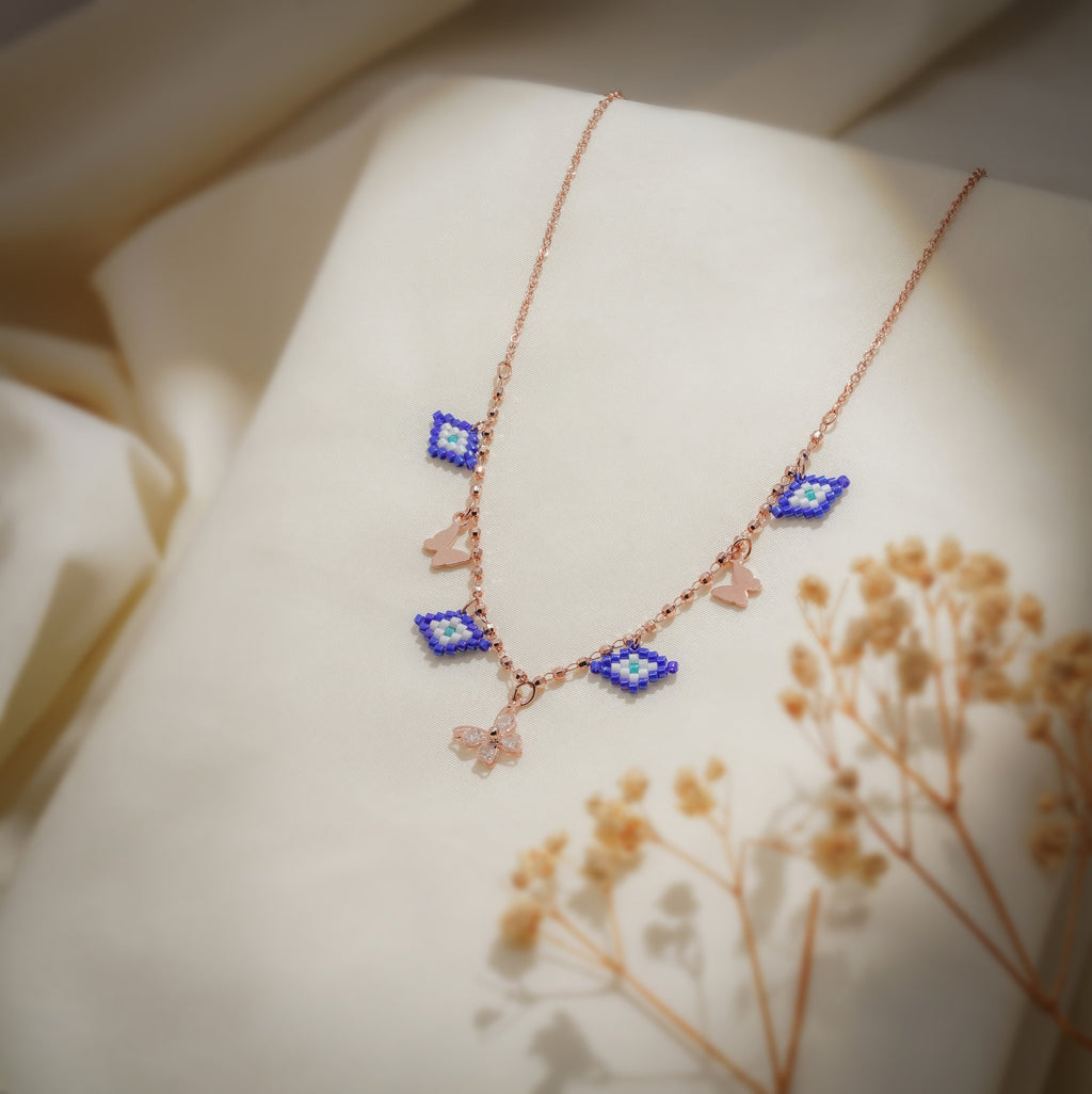 Rose-gold charm necklace with cobalt beaded evil-eye charms and butterfly charms on cream fabric