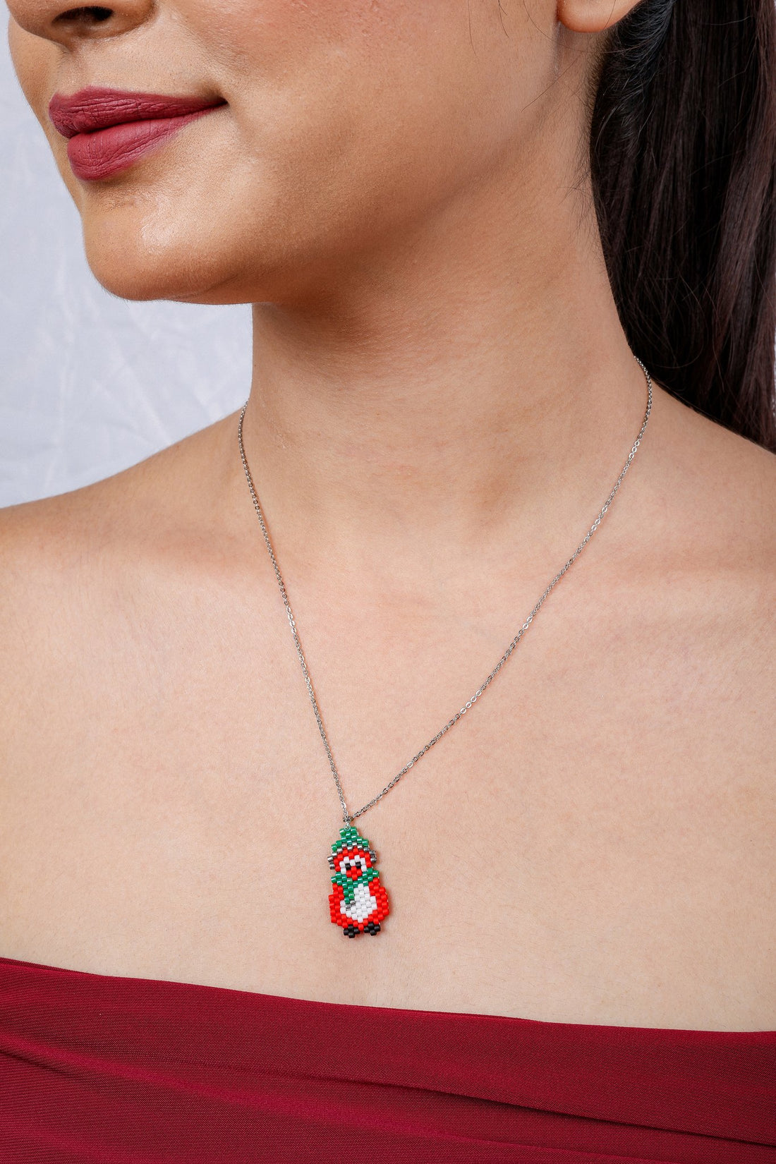 Model wearing silver chain with beaded red and white penguin pendant in a green hat and scarf