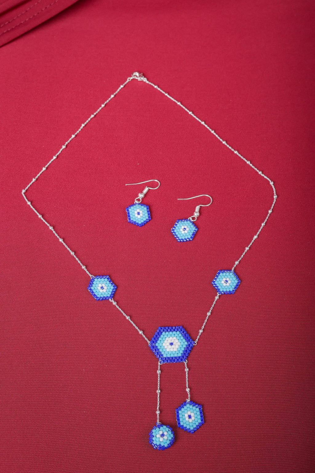 Silver evil eye necklace with five beaded blue hexagons and matching hook earrings on red fabric