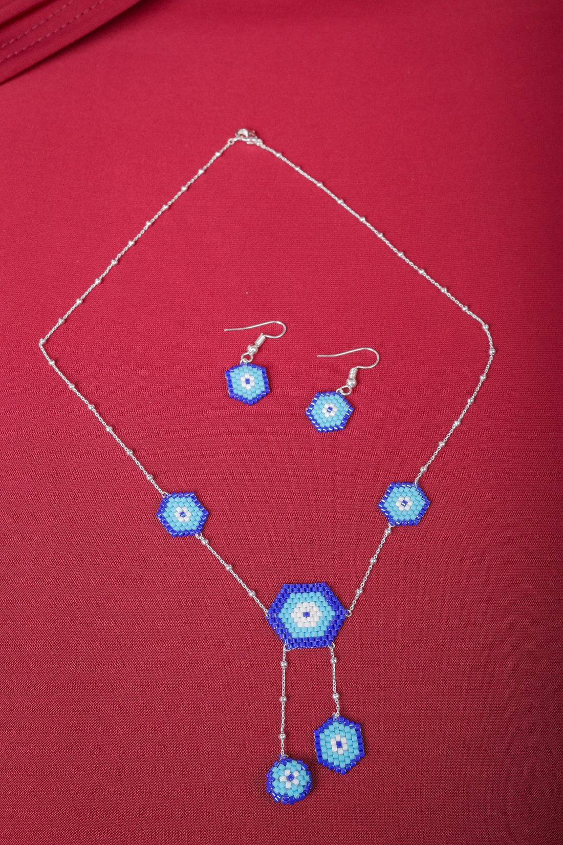 Silver evil eye necklace with five beaded blue hexagons and matching hook earrings on red fabric
