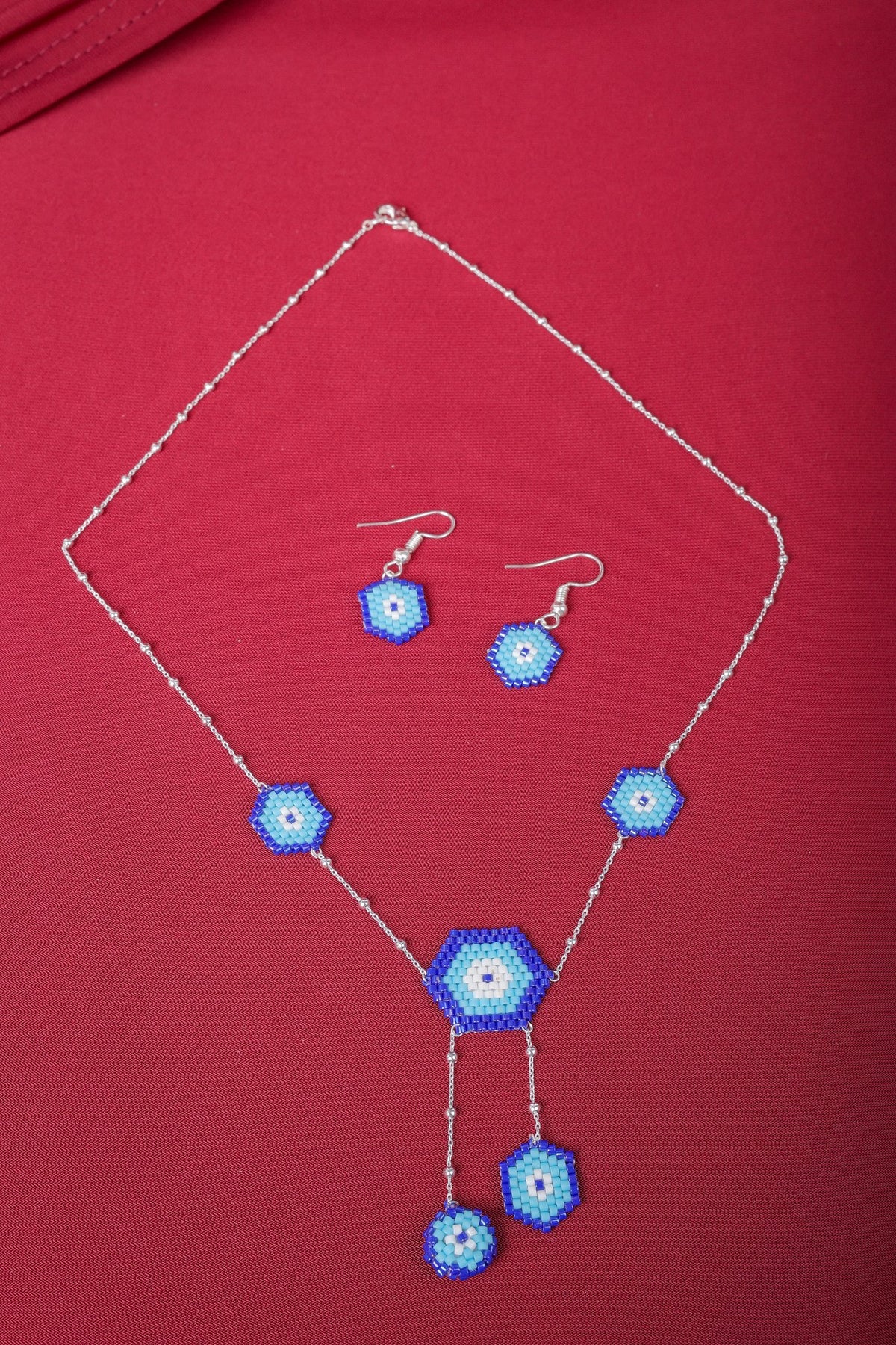 Silver evil eye necklace with five beaded blue hexagons and matching hook earrings on red fabric