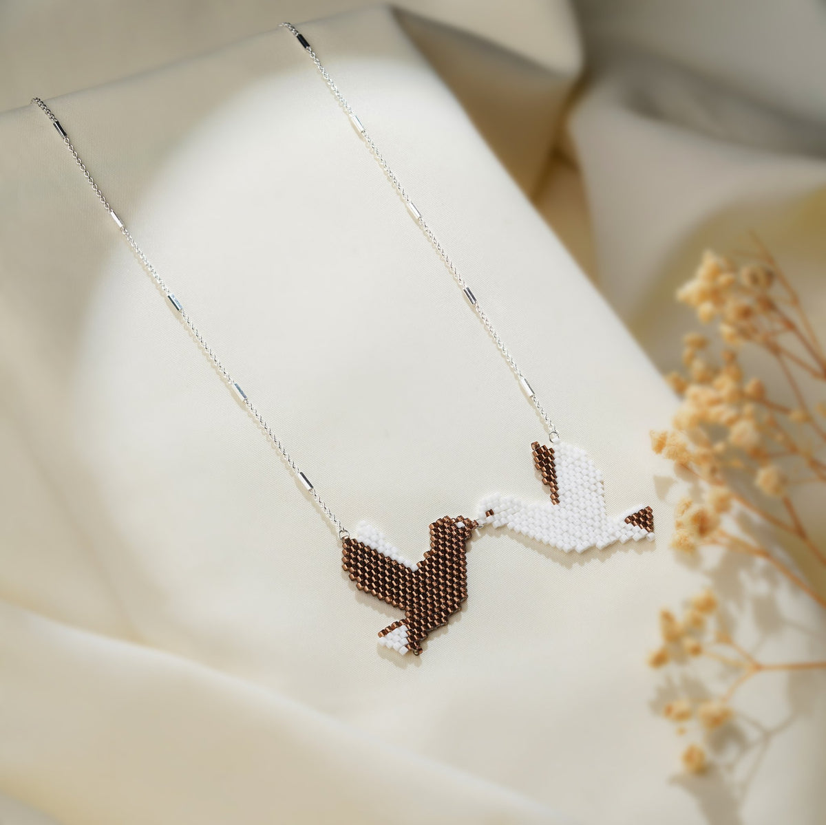 Brown and white beaded kissing doves pendant on a silver chain beside dried flowers