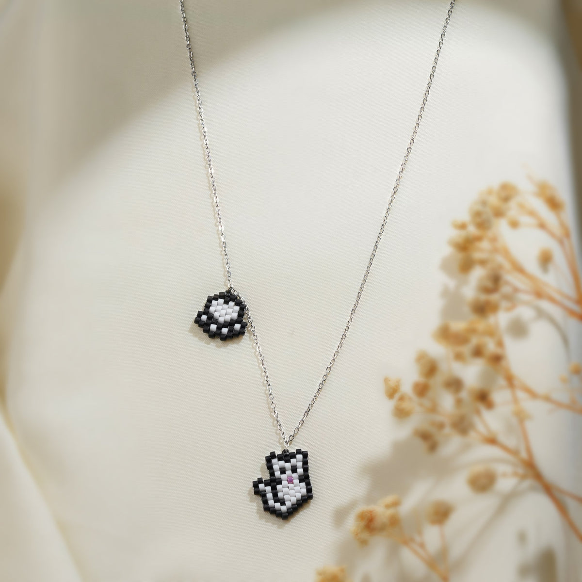 Silver chain necklace with white pixel-beaded cat charm and black and white paw-print charm on cream