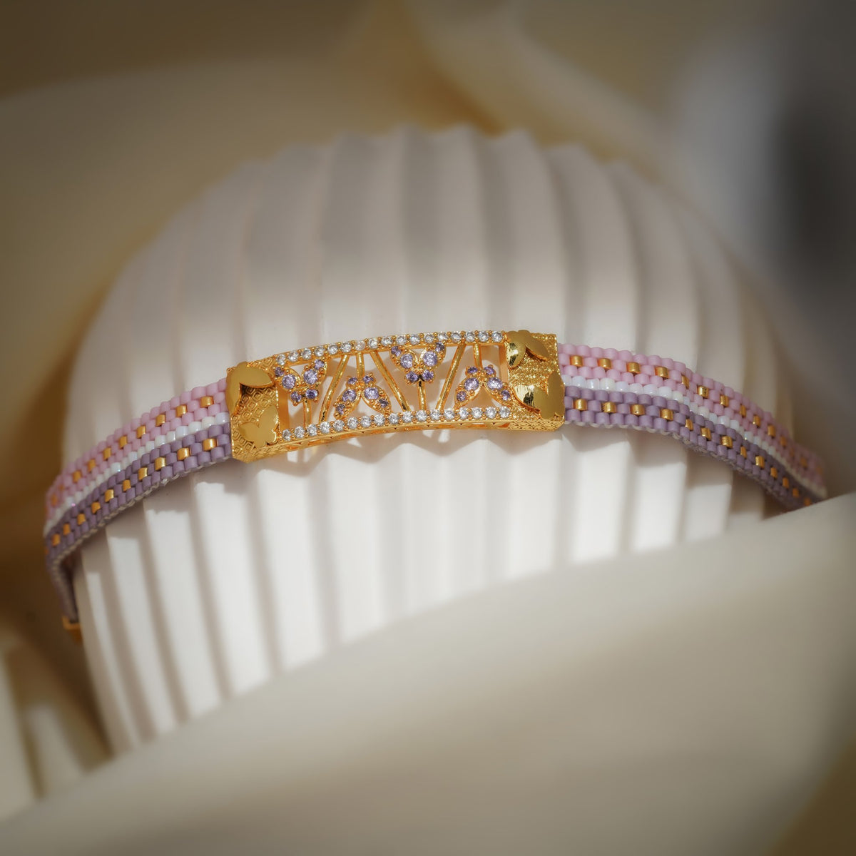 Lilac, pink and white woven bead bracelet with gold crystal-set centre plate on a ribbed stand