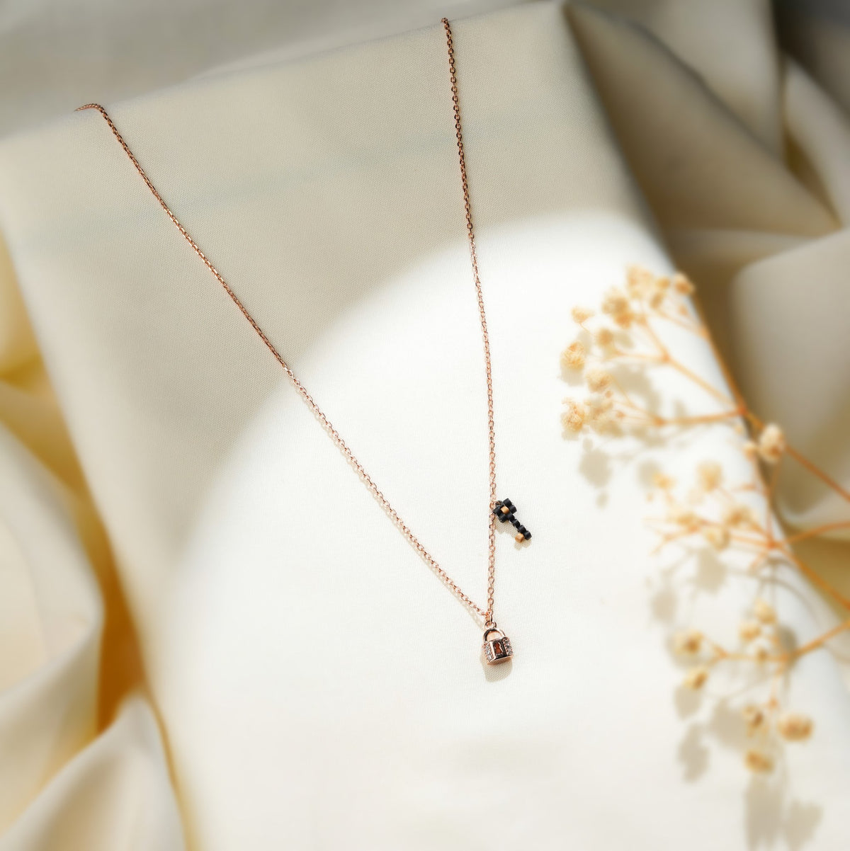 Rose-gold-tone lock and key necklace draped on cream fabric beside dried gypsophila