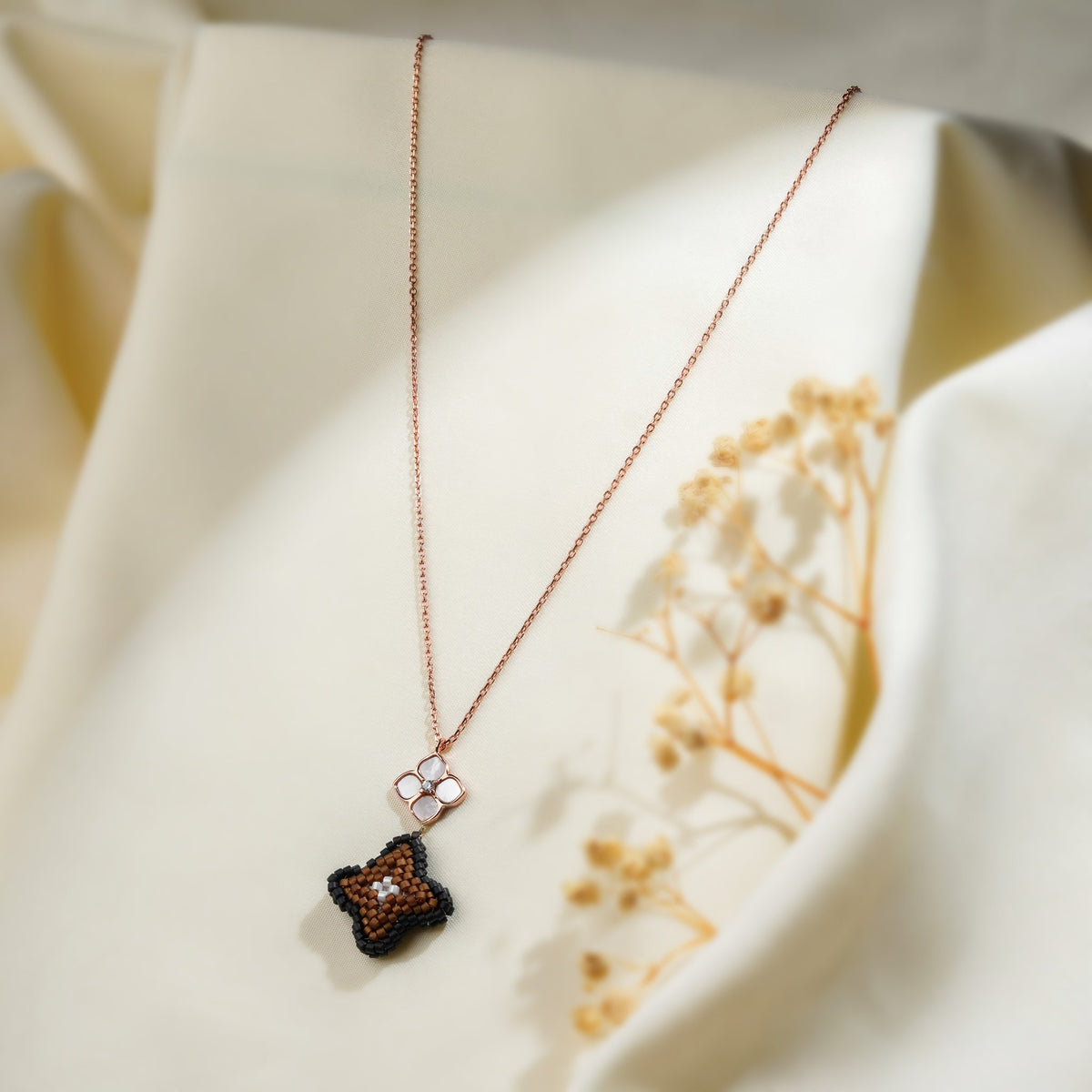 Rose-gold necklace with pearly four-petal flower and black and brown beaded clover pendant on cream fabric