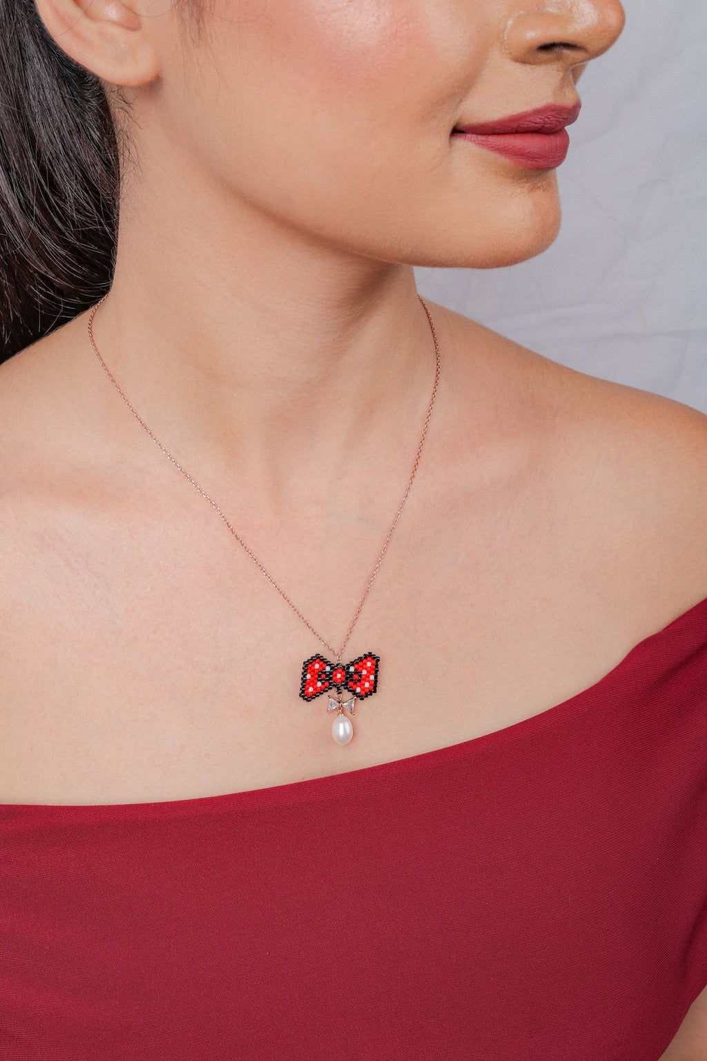 Model wearing the red beaded bow pendant with pearly drop on a rose-gold chain, side view