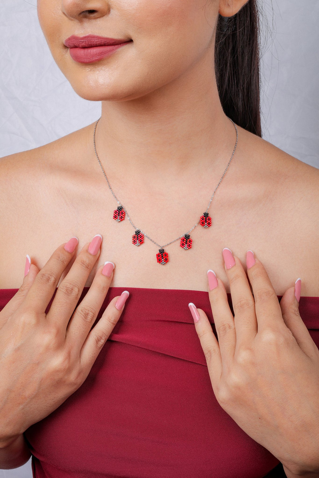 Model in a red off-shoulder top wearing a ladybug charm necklace, hands at chest