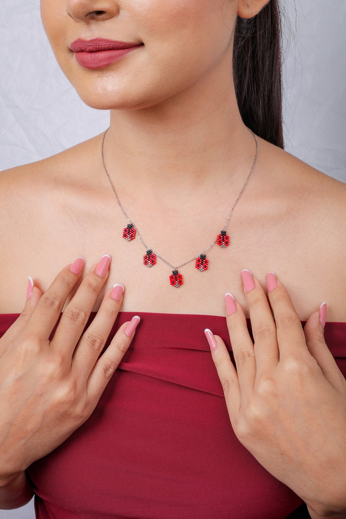 Model in a red off-shoulder top wearing a ladybug charm necklace, hands at chest