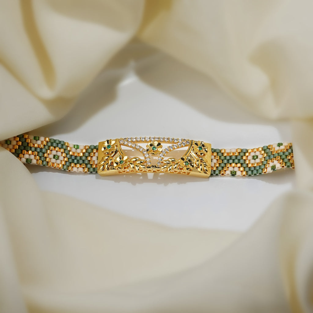 Woven sage green, gold and white bead bracelet with gold floral centre plate laid flat on cream silk