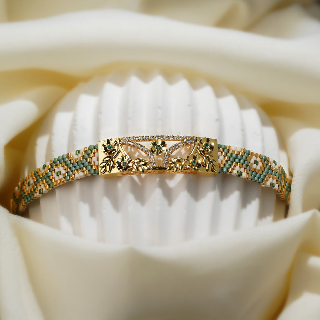 Gold floral plate and zigzag bead pattern of the sage green woven cuff on a white stand