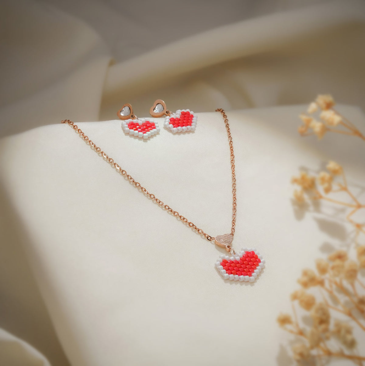 Red beaded heart necklace and earring set laid on cream fabric beside dried flowers