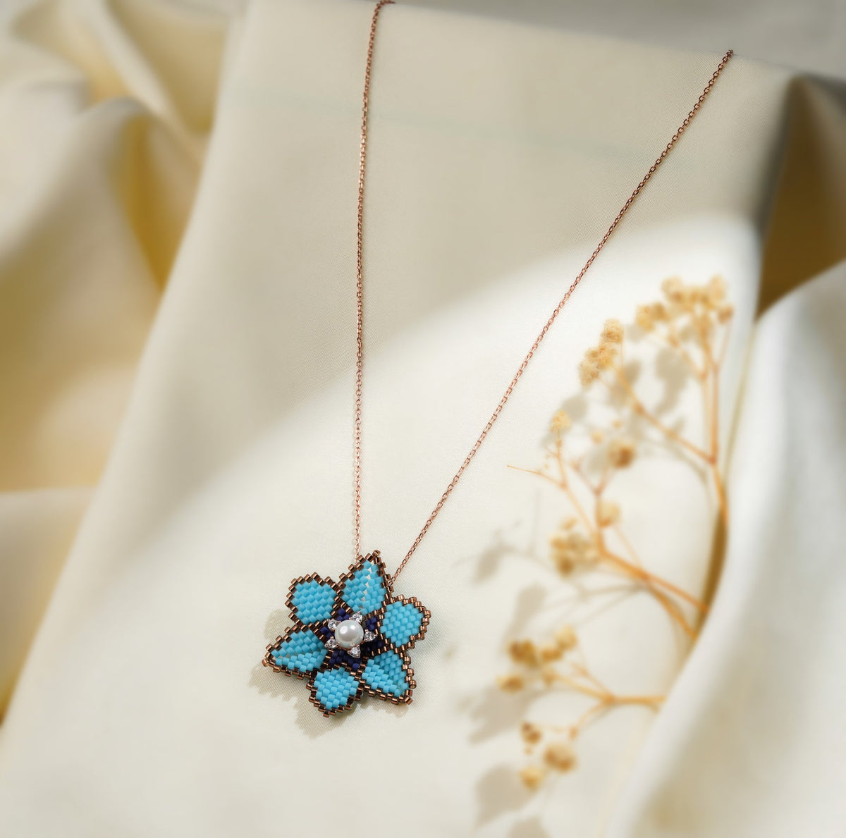 Turquoise beaded flower pendant on a rose-gold-tone chain draped over cream fabric