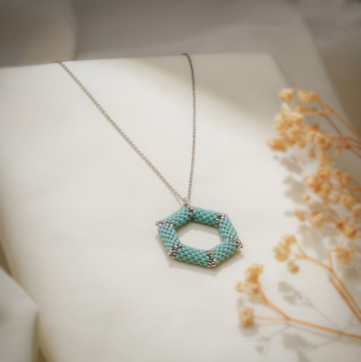 Open turquoise beaded hexagon pendant on a fine silver-tone chain over cream fabric